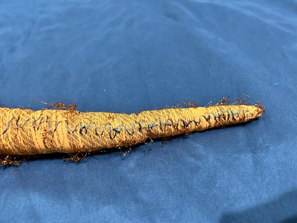 A fabric tentacle made of orange fabric with shiny metallic thread through it. Some of the thread is coming off. There is a zigzag line of conductive thread so along the side of it. Background is blue fabric.
