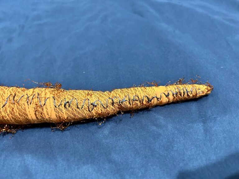 A fabric tentacle made of orange fabric with shiny metallic thread through it. Some of the thread is coming off. There is a zigzag line of conductive thread so along the side of it. Background is blue fabric.